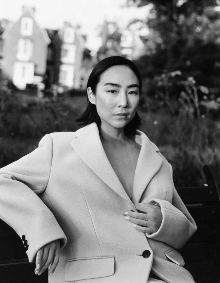 Monochrome portrait of an Asian woman wearing a blazer sitting on a park bench, with trees and buildings visible in the background.