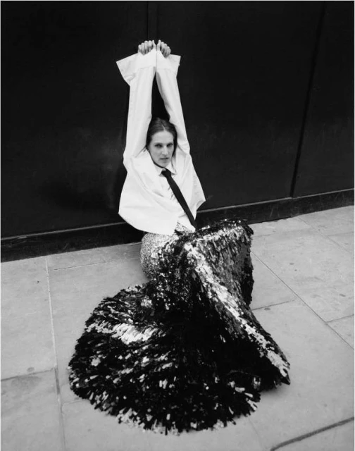 A woman sits on a floor in a white dress shirt, black tie, and sparkly skirt, her hands raised up over her head. Black background.