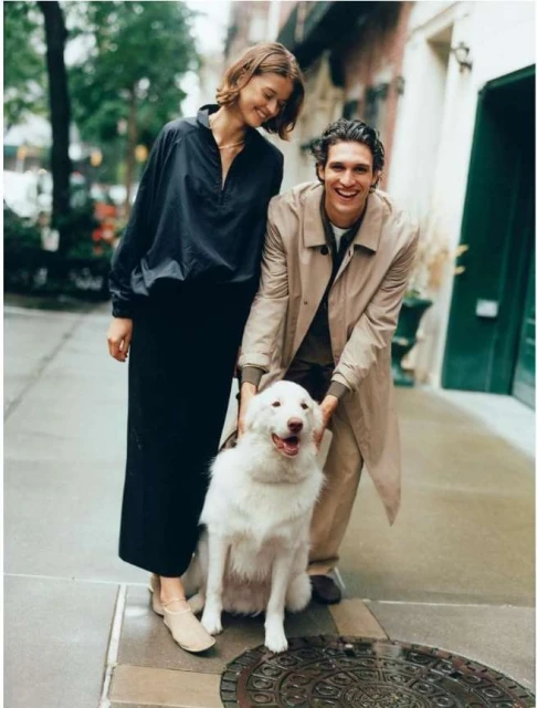 Smiling couple walking a white dog along a city street with brick buildings and green trees. The dog sits looking at the camera.