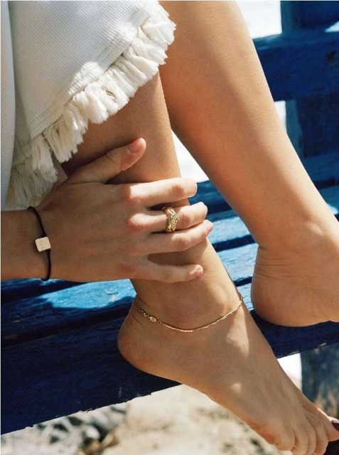 Close-up of an adult woman's leg and foot adorned with a gold anklet, along with her hand featuring a gold ring, set against a blue background.