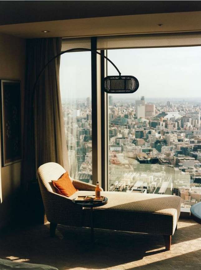 Interior view of a neutral-toned chaise lounge with orange pillow and lamp, set against a backdrop of a sprawling city view.