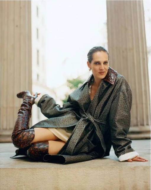 A woman in a gray coat with polka dots poses on a stone step, her leg raised in a knee-high snakeskin boot, shot against a blurred background.
