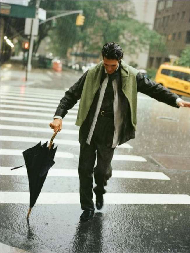 Man walking in the rain across a crosswalk with an umbrella, wearing a green vest, dark suit, and black shoes in a city street.