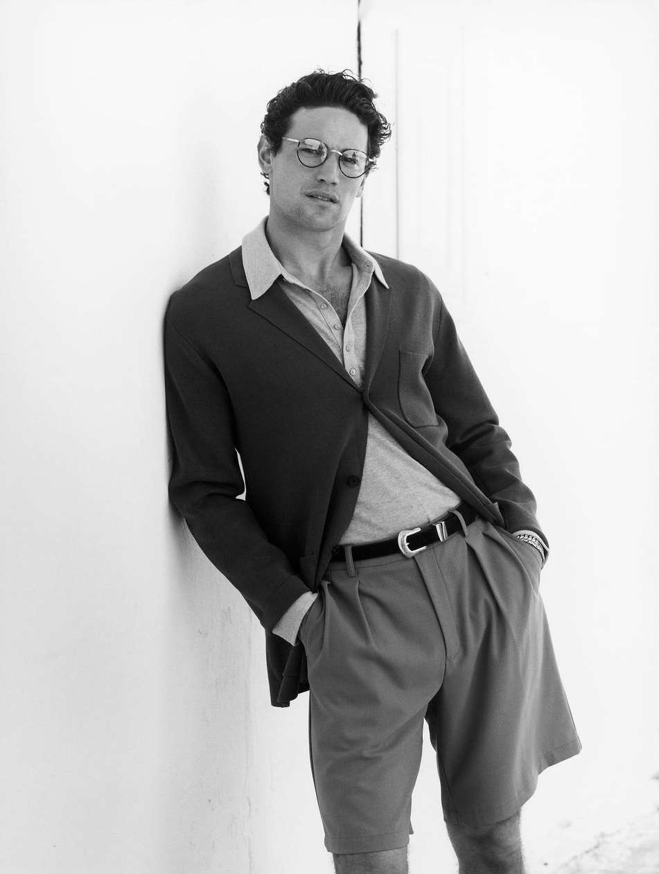 Black and white portrait of a man in a blazer, shorts, and glasses leaning against a wall with his hands in his pockets.