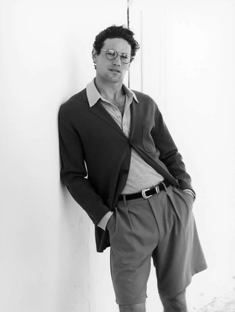 Black and white portrait of a man in a blazer, shorts, and glasses leaning against a wall with his hands in his pockets.