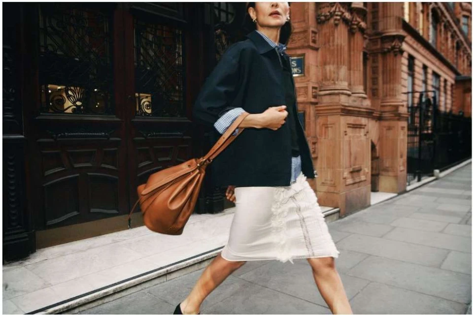Elegant woman in navy jacket and skirt carrying tan leather handbag on a city sidewalk in front of architectural building.
