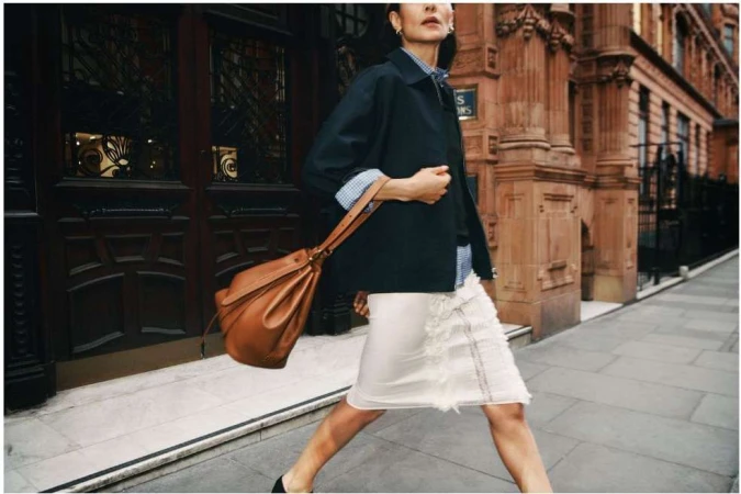Elegant woman in navy jacket and skirt carrying tan leather handbag on a city sidewalk in front of architectural building.