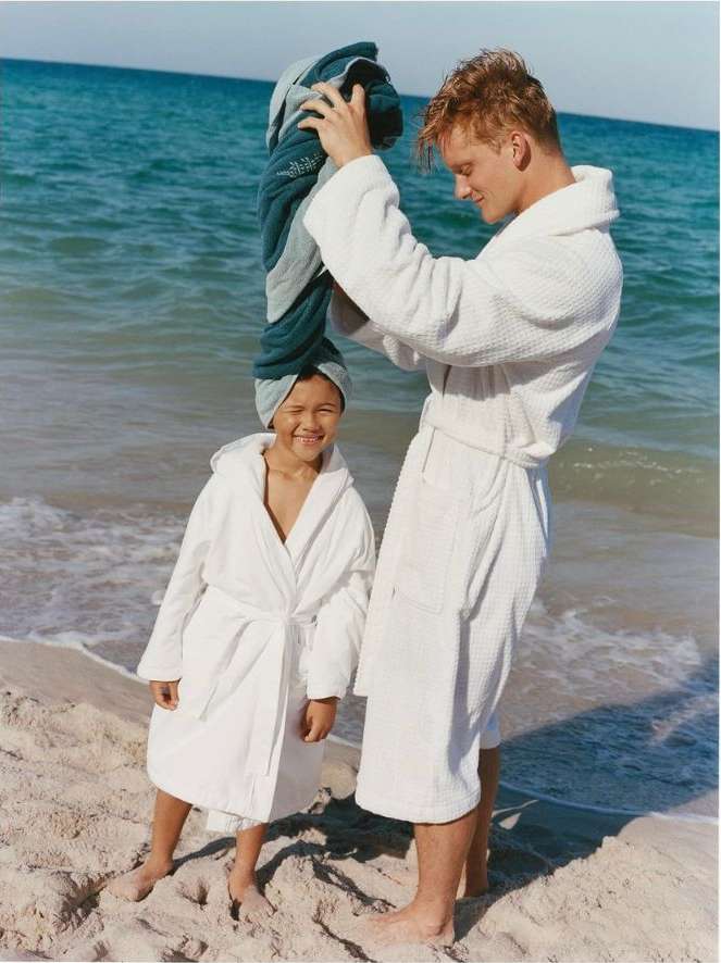 Father and daughter in white robes stand on a beach next to the ocean. The father is placing a blue towel on the child's head.