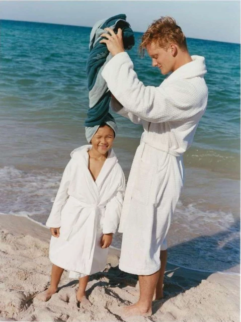 Father and daughter in white robes stand on a beach next to the ocean. The father is placing a blue towel on the child's head.
