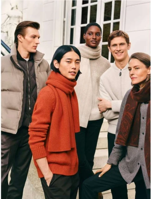Five people in stylish, casual clothing stand outdoors in front of a building, posing for a fashion advertisement. They wear sweaters and scarves.