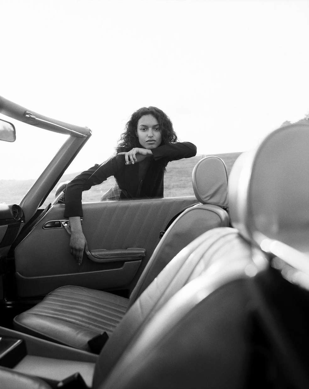 Black and white portrait of a woman in a jacket leaning against a classic convertible car, captured in an outdoor setting.