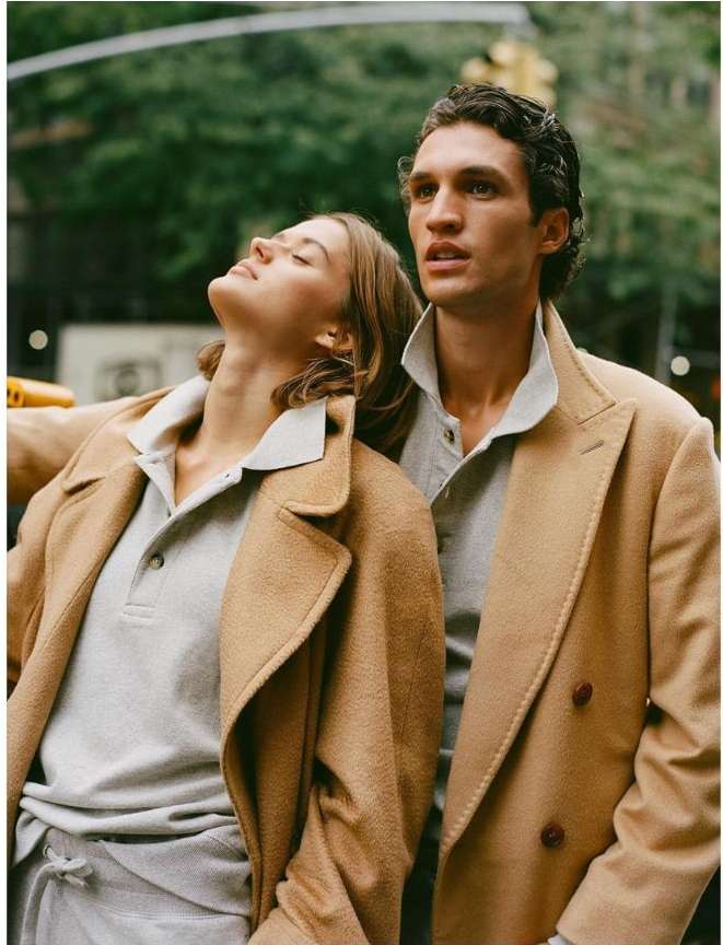 A stylish couple standing closely outdoors, both wearing tan overcoats and grey polo shirts, with a blurred urban backdrop.