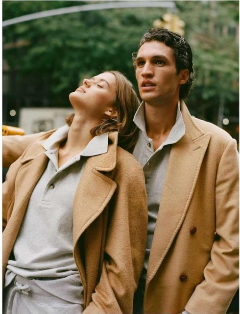 A stylish couple standing closely outdoors, both wearing tan overcoats and grey polo shirts, with a blurred urban backdrop.