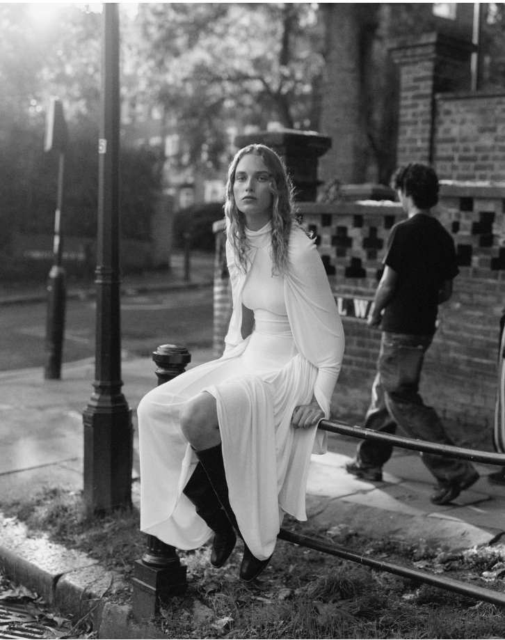 A woman in a long white dress and black boots sits on a metal guardrail in a black and white outdoor street scene.