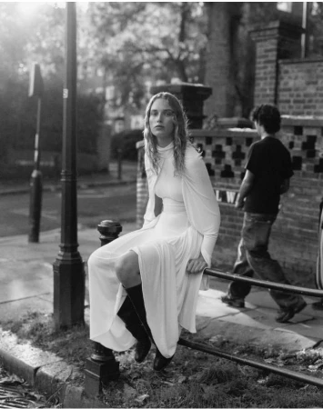 A woman in a long white dress and black boots sits on a metal guardrail in a black and white outdoor street scene.
