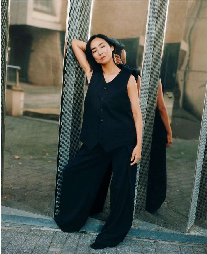 Adult female in a sleeveless black top and wide-leg pants leans against a modern mirrored wall outdoors in an urban setting.