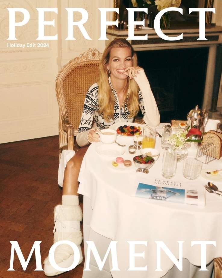 Smiling woman with long blonde hair poses at a breakfast table set with fresh fruit, pastries, juice, and hot beverages in an interior setting.