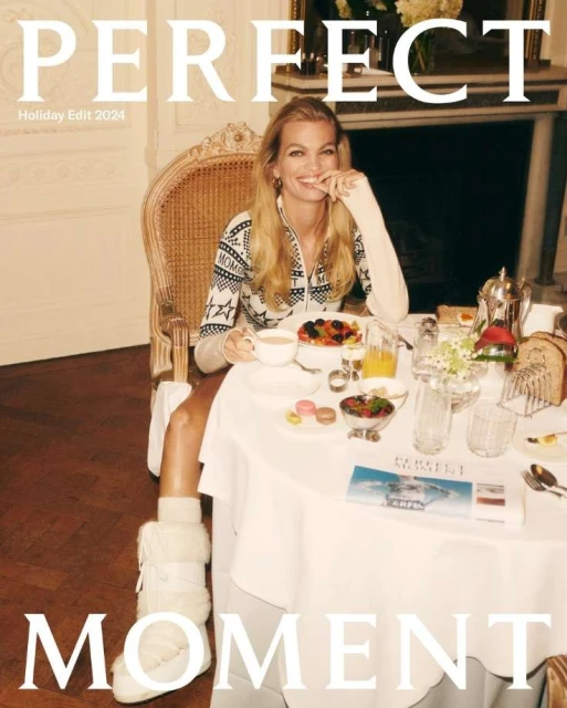 Smiling woman with long blonde hair poses at a breakfast table set with fresh fruit, pastries, juice, and hot beverages in an interior setting.