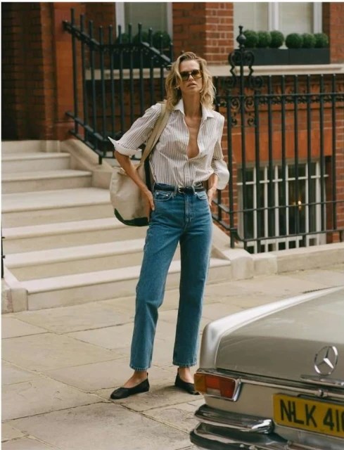 A slender adult woman stands on a sidewalk in front of a brick building wearing jeans, a striped shirt, and sunglasses with a shoulder bag.