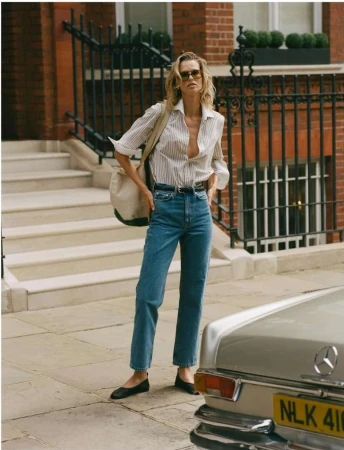 A slender adult woman stands on a sidewalk in front of a brick building wearing jeans, a striped shirt, and sunglasses with a shoulder bag.
