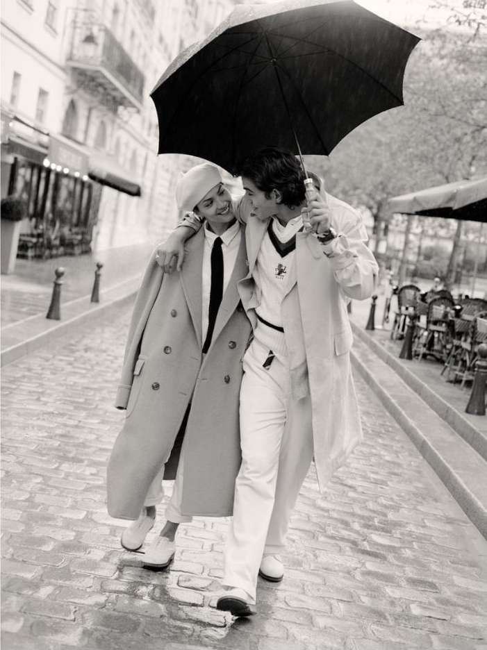 Black and white photograph of a stylish couple walking under an umbrella on a cobblestone street, surrounded by buildings and outdoor cafes.