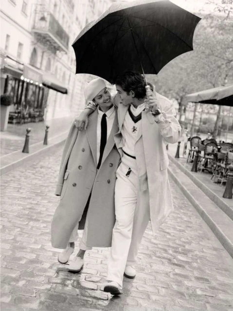 Black and white photograph of a stylish couple walking under an umbrella on a cobblestone street, surrounded by buildings and outdoor cafes.