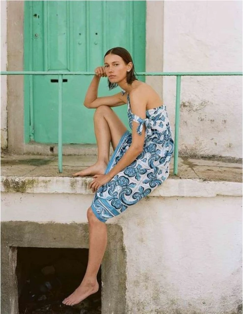 Woman in blue and white sarong sits barefoot on ledge in front of turquoise door. Natural light, outdoor setting, fashion shot.
