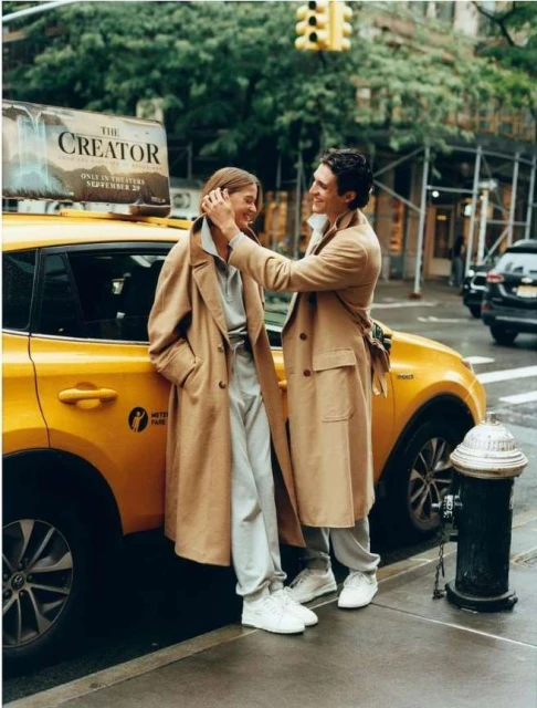 Couple in beige trench coats pose by a yellow taxi cab on a city street with surrounding buildings and a vintage fire hydrant.