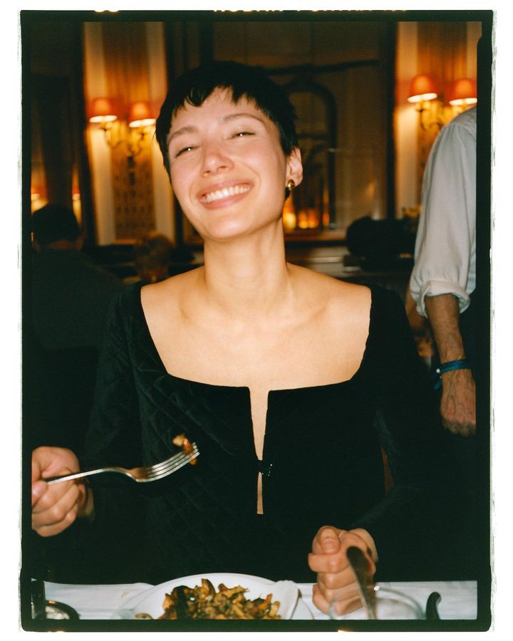 Smiling woman in a black dress holds a fork and knife at a restaurant table with plate of food and red lamps in the background.