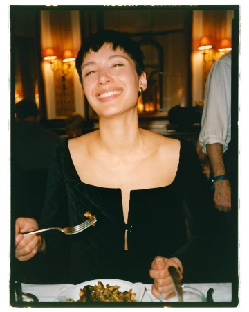 Smiling woman in a black dress holds a fork and knife at a restaurant table with plate of food and red lamps in the background.