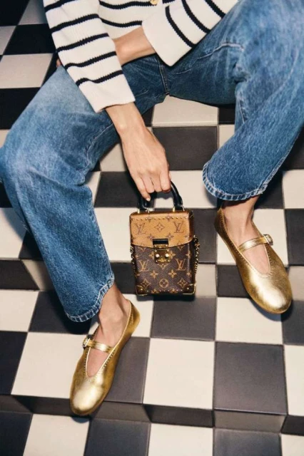 A woman sits on a black-and-white checkered surface, wearing jeans, a striped shirt, and gold shoes, while holding a luxury purse.