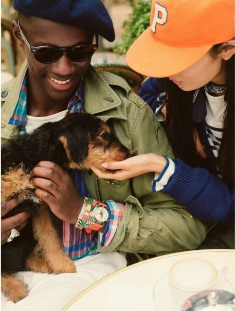 A black dog is the central focus as a smiling African American man and an Asian woman with an orange cap pet the dog.