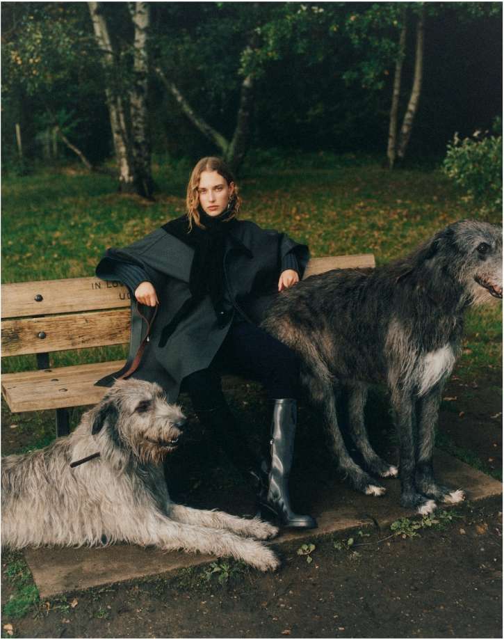 Woman in a dark coat sits on a wooden bench in a park with two large Irish Wolfhound dogs, creating a fashion editorial scene.
