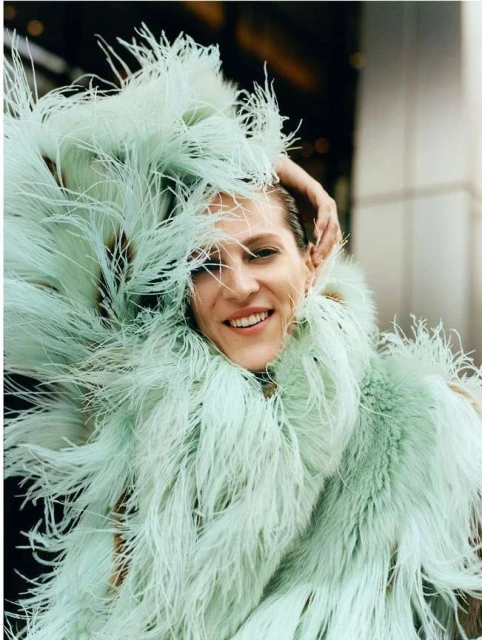 Smiling woman in a bright-green feather boa poses with her hand on her head in front of a neutral building on a sunny day.