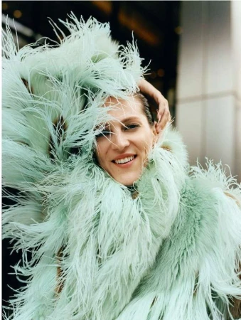Smiling woman in a bright-green feather boa poses with her hand on her head in front of a neutral building on a sunny day.