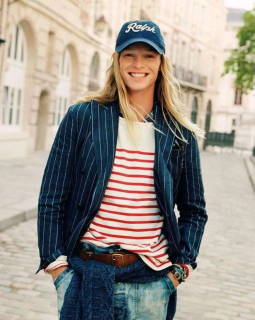 Smiling blond man in striped blazer and baseball cap. He wears jeans and stands in an outdoor urban setting with light stone buildings.
