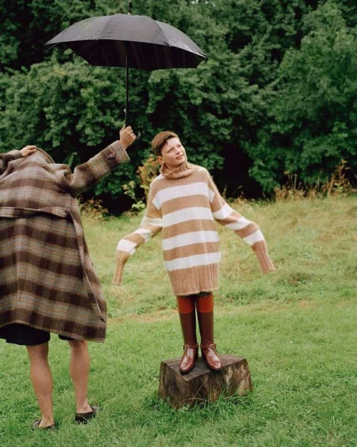 Young boy stands on a tree stump in a field, wearing a striped sweater and boots, while an adult holds an umbrella over him.