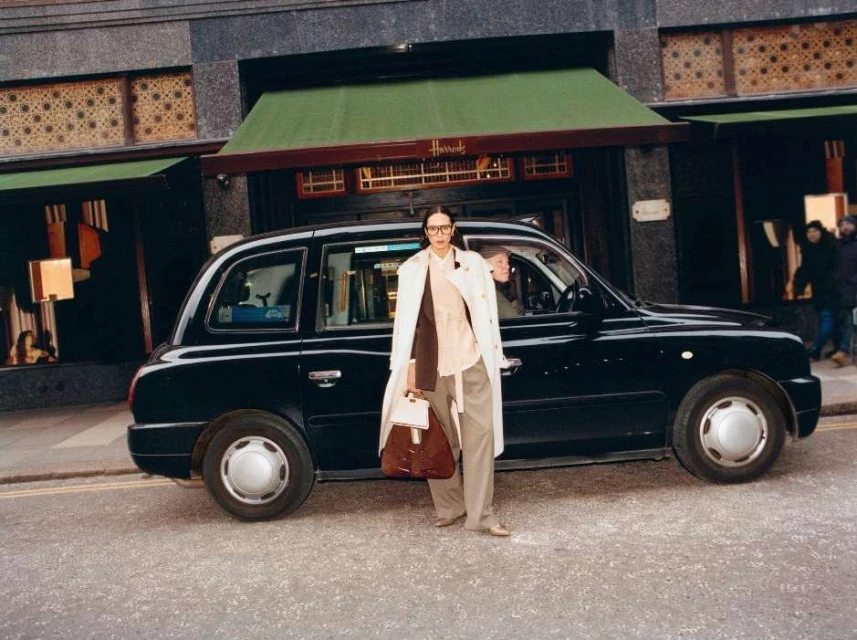 An adult woman in a trench coat poses next to a black taxi cab with the Harrods department store visible in the background in London.