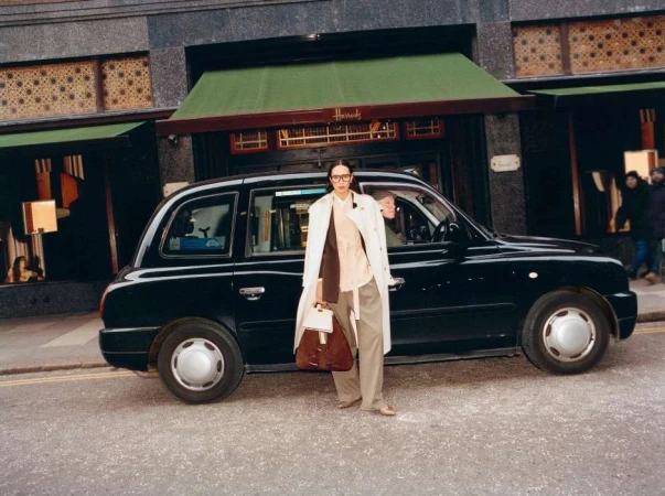 An adult woman in a trench coat poses next to a black taxi cab with the Harrods department store visible in the background in London.