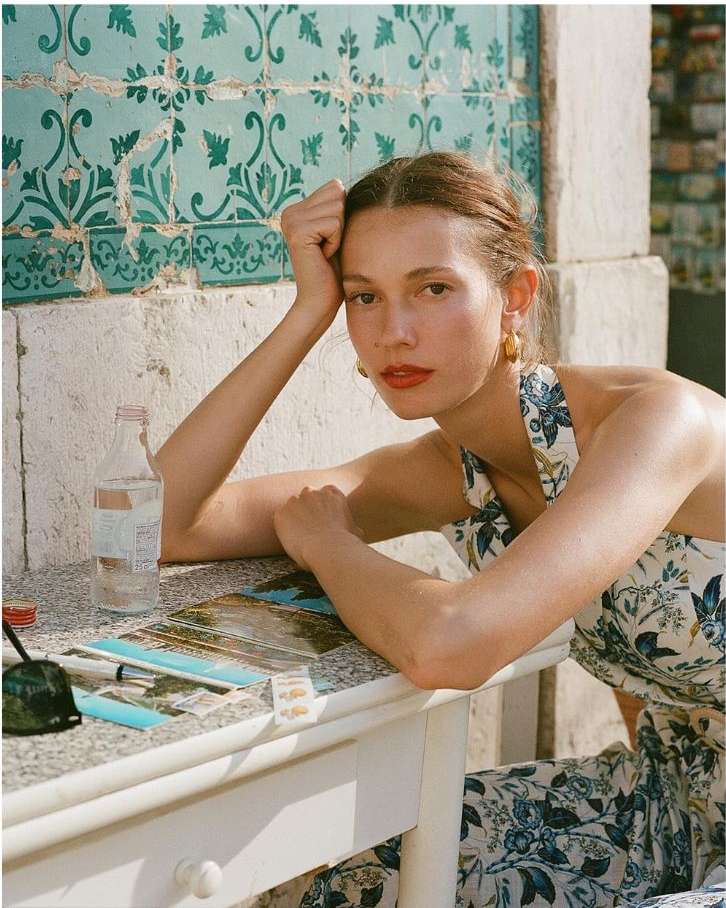 Young woman in floral dress at outdoor cafe with water bottle, sunglasses, postcards on table, leaning on arm against tile wall.