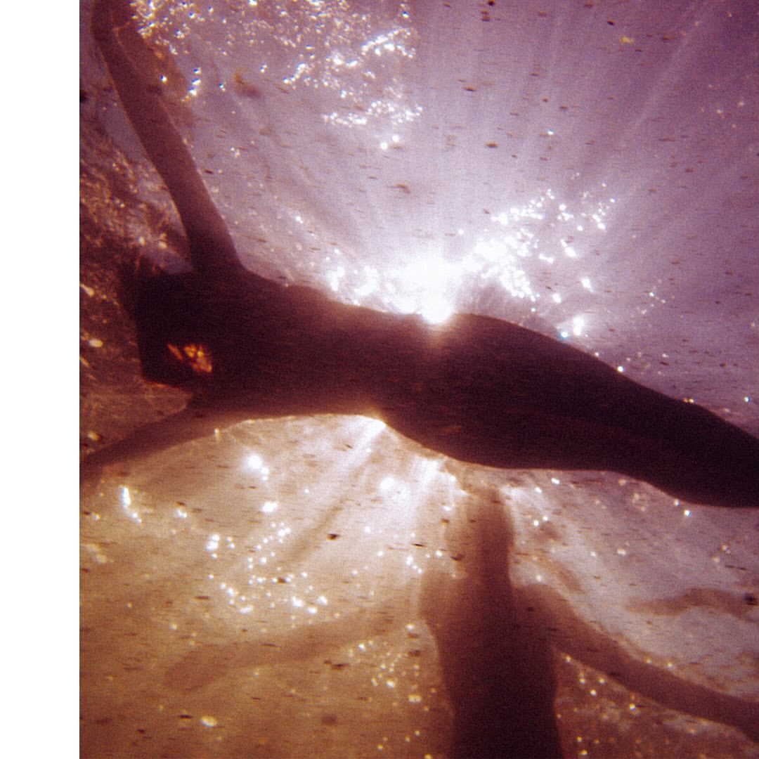 Underwater view of a silhouetted adult woman swimming, illuminated by sunbeams piercing through the water's surface, creating a serene aquatic scene.