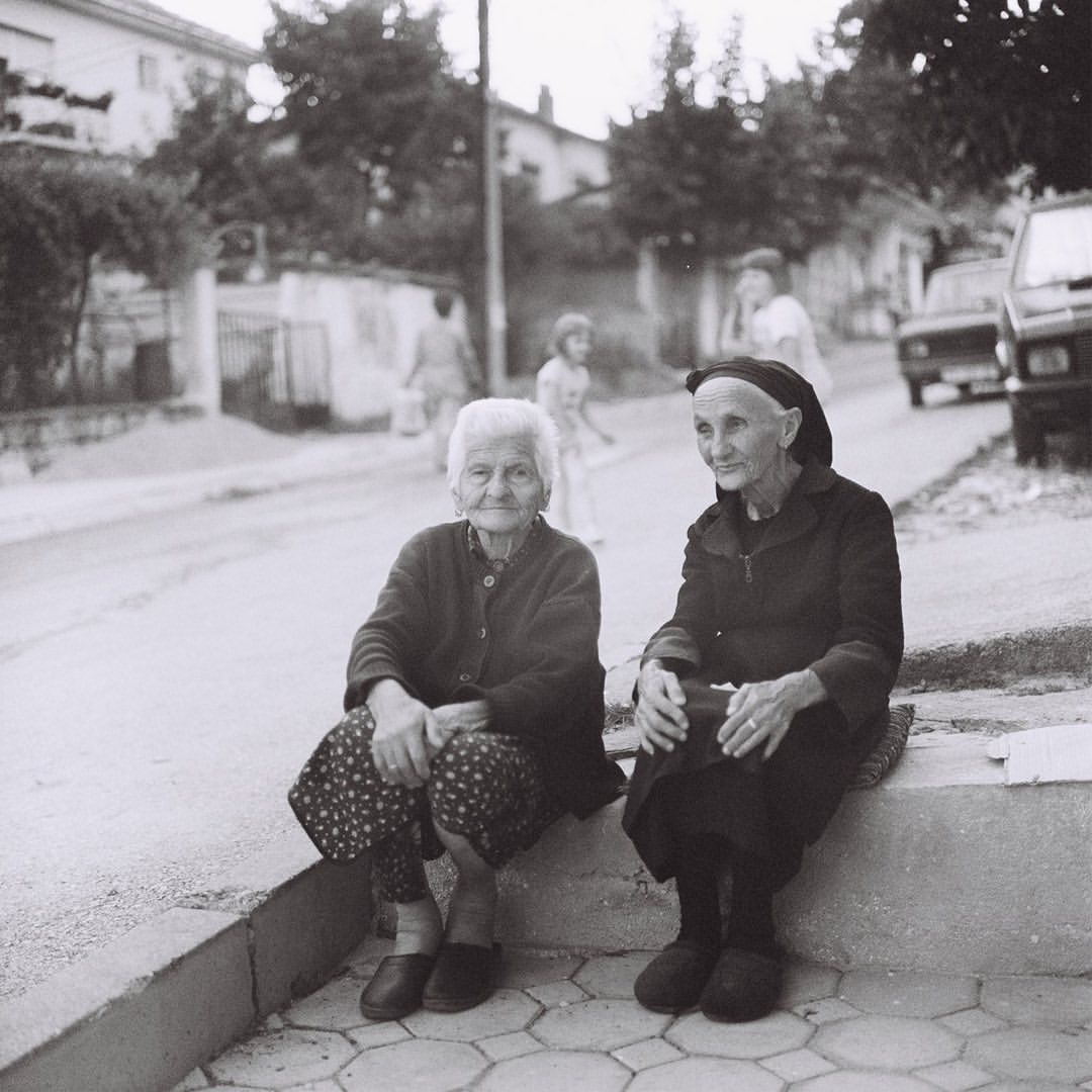 Two elderly women are sitting on a curb in a monochromatic outdoor shot. A street scene with pedestrians, buildings, and a car completes the image.