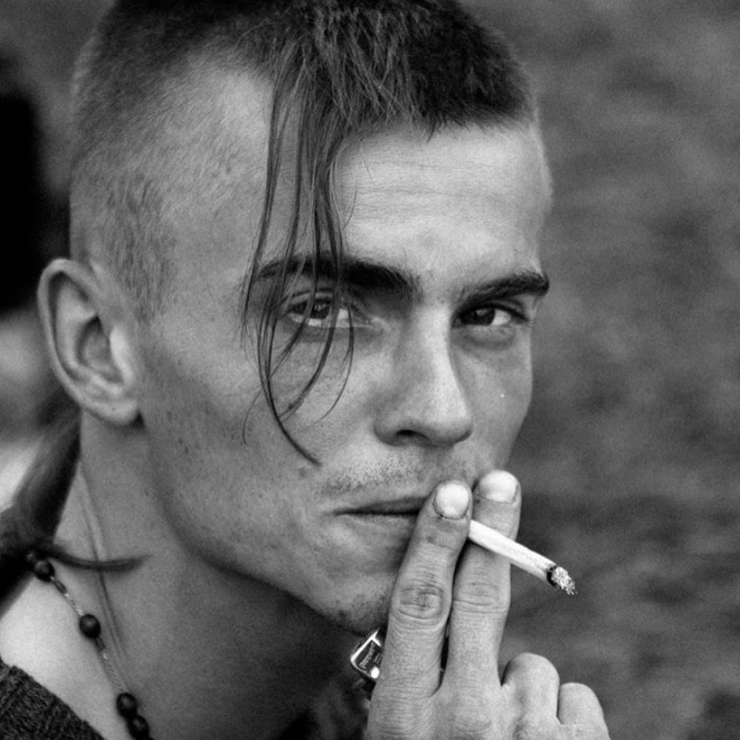 Monochrome portrait of a young adult man smoking a cigarette, featuring a distinctive hairstyle and casual attire in an outdoor setting.