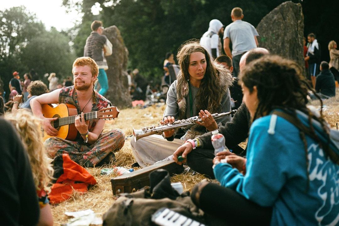 Outdoor music scene with a man playing guitar and a woman with a flute among a seated group of people on grass in a natural environment.