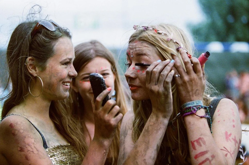 Three women with body paint apply makeup at an outdoor event. One woman holds a mascara wand to her eye, another holds a phone.