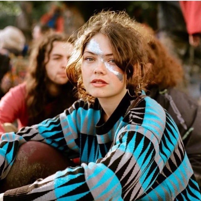 Portrait of a young woman with glitter makeup, wearing a patterned sweater, surrounded by a blurred group of people outdoors.