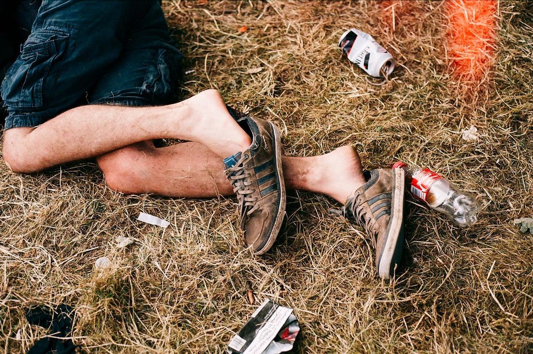 A man lies on dry grass wearing sneakers, with trash scattered nearby, creating a scene of post-event relaxation or litter.