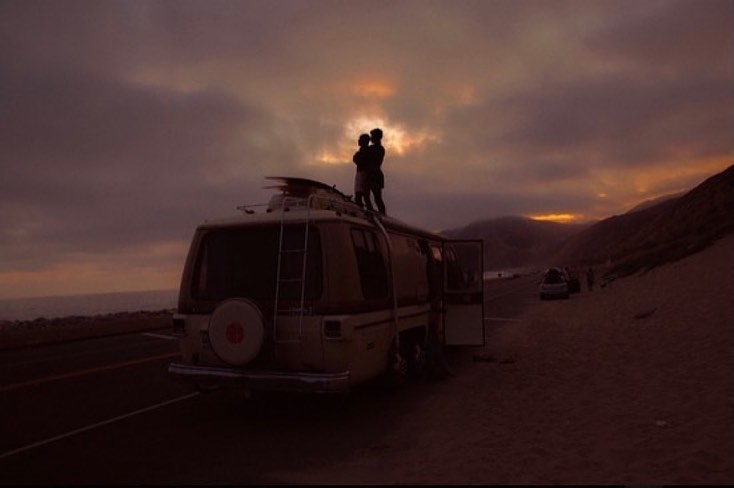 Couple stands atop camper van parked by the beach at sunset. Dramatic cloudy sky and coastal mountain range form the backdrop.
