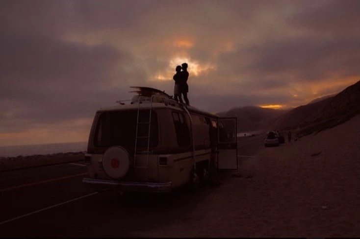 Couple stands atop camper van parked by the beach at sunset. Dramatic cloudy sky and coastal mountain range form the backdrop.