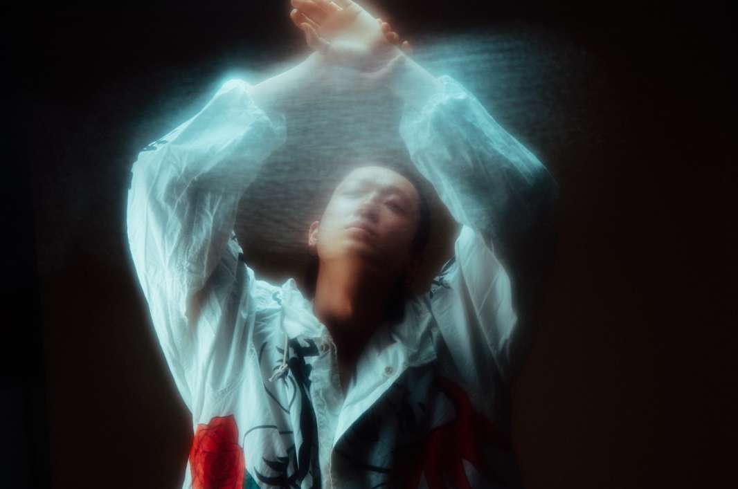 Woman in white floral jacket raises her arms in a blurred motion against a dark background, artistic and dreamy.